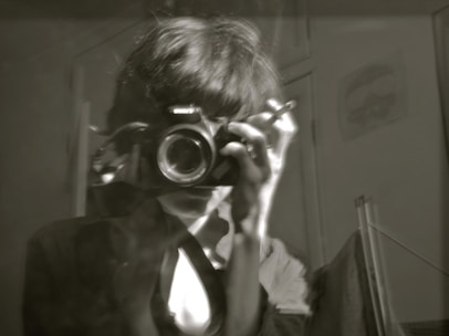 A black and white image of a person holding a camera up to their face, possibly taking a photograph. Their hair is slightly messy, and they are wearing a jacket over a shirt. The room is dimly lit, and the background shows part of a wall with a faint poster or picture.