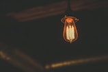 An Edison-style light bulb glowing warmly in a dimly lit environment. The filament is clearly visible inside the bulb, casting a soft amber light around it.