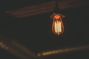 An Edison-style light bulb glowing warmly in a dimly lit environment. The filament is clearly visible inside the bulb, casting a soft amber light around it.