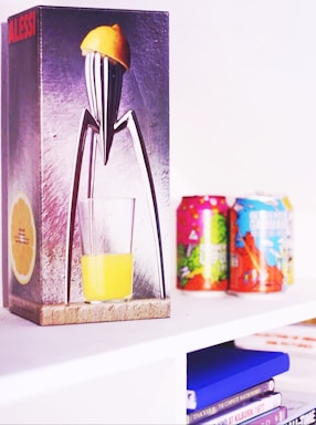 A sleek and modern citrus juicer with a lemon on top is depicted on the packaging of an Alessi product. Beside it are colorful cans on a white shelf, and below are stacks of books and DVDs.