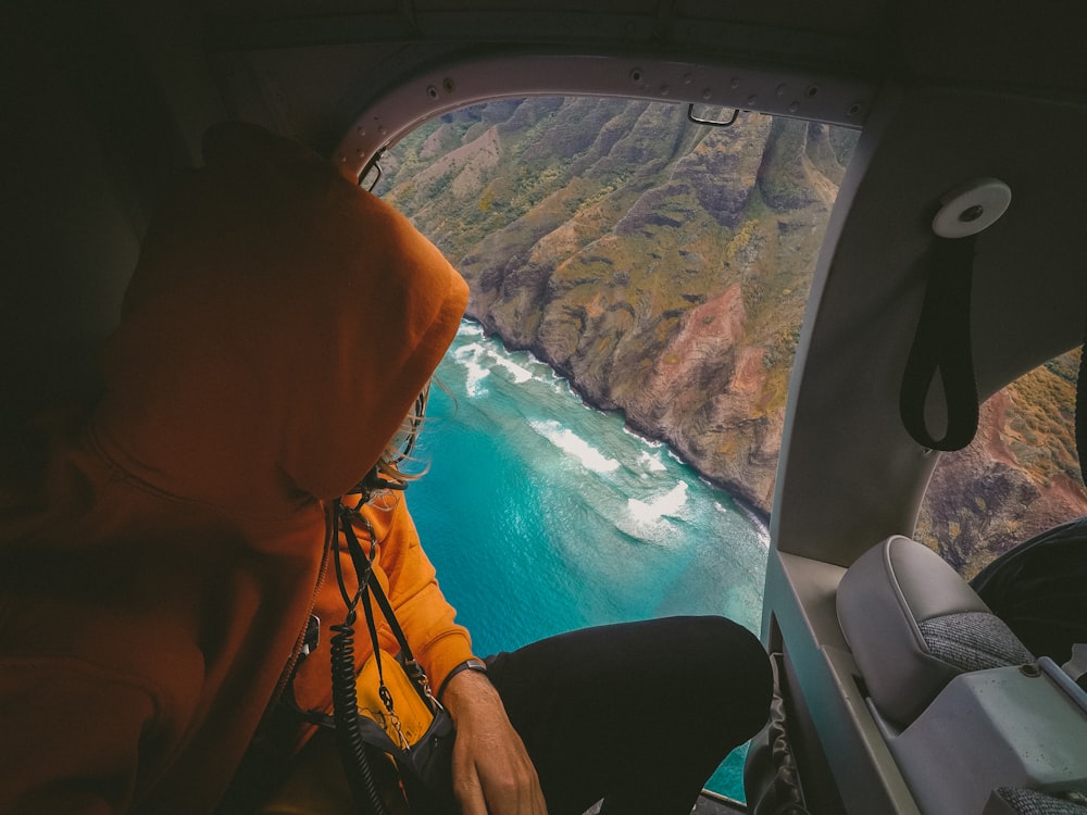A person in an orange parka sitting in a helicopter with an azure coast visible in the window