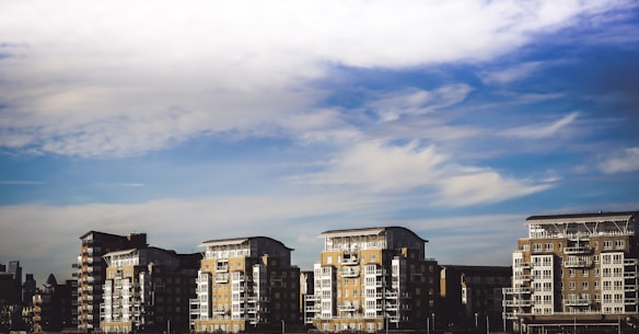 Several modern apartment buildings with numerous windows and balconies stand under a vast, partly cloudy sky. The architecture reflects urban residential structures, featuring muted colors that blend harmoniously with the environment.