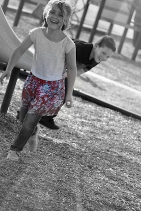 A young girl in colorful floral shorts runs joyfully, with another child playfully peeking from behind a playground slide. The background is slightly blurred, focusing attention on the children's expressions and movement.