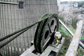 A large, industrial pulley system is mounted on a platform with thick green cables running over it, showing signs of rust and patina. Surrounding the pulley, there are industrial structures, a concrete cooling tower, and metal scaffolding. The background includes trees and an overcast sky.