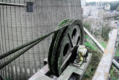 A large, industrial pulley system is mounted on a platform with thick green cables running over it, showing signs of rust and patina. Surrounding the pulley, there are industrial structures, a concrete cooling tower, and metal scaffolding. The background includes trees and an overcast sky.