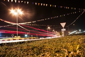 A nighttime traffic scene with light trails from vehicles passing on the road. Streetlights illuminate the area, casting bright, star-like flares. Colorful triangular flags are strung across the scene, adding a festive touch. A traffic sign and some greenery are visible in the foreground.