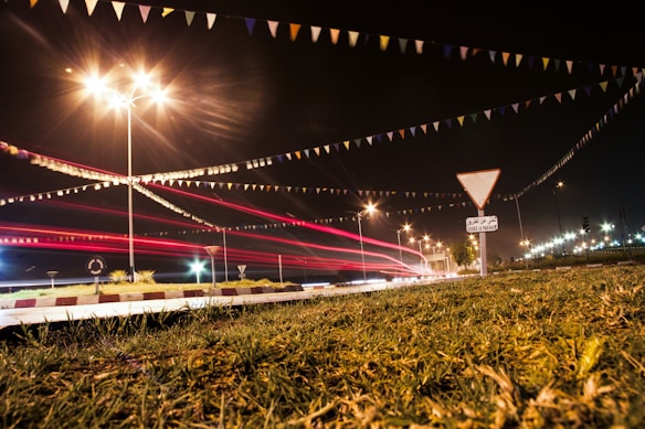 A nighttime traffic scene with light trails from vehicles passing on the road. Streetlights illuminate the area, casting bright, star-like flares. Colorful triangular flags are strung across the scene, adding a festive touch. A traffic sign and some greenery are visible in the foreground.