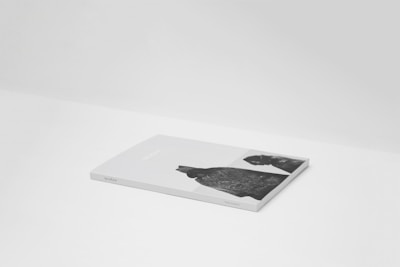 A flat, closed book or magazine with a light gray cover, placed on a white surface. The cover features black and white photographs or illustrations and some text.