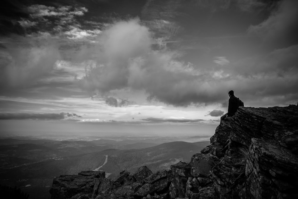 Fotografia em tons de cinza do homem sentado no pico da montanha sob nuvens cumulus