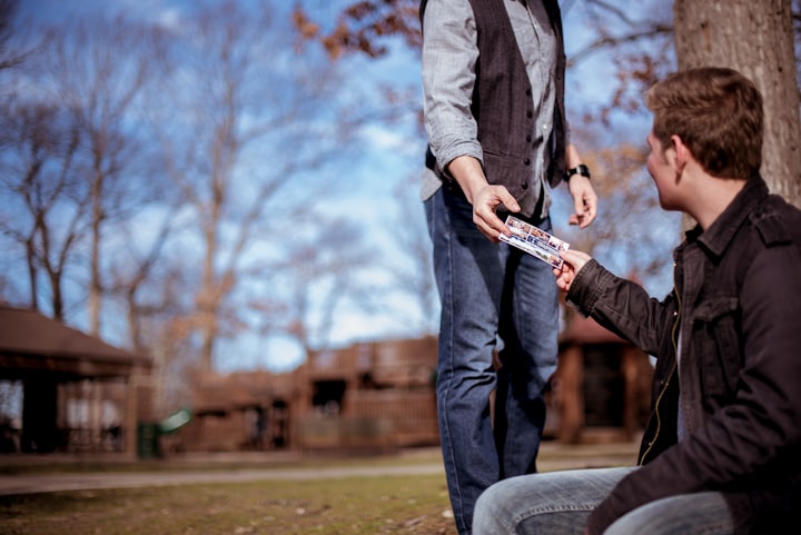 man handing another man a note, likely some kind of referral in a public park in the fall by a tree 