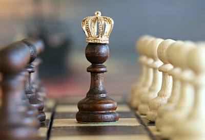 A dark chess pawn wearing a golden crown stands prominently in the center of a chessboard, flanked by rows of white and dark pawns. The chess pieces are aligned neatly, creating a symmetrical and orderly appearance.