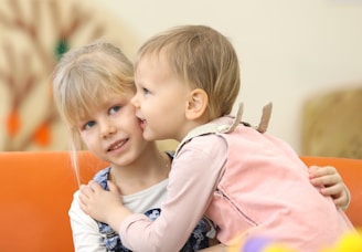 Two young children are interacting on an orange sofa. One child, wearing a pink outfit, is whispering in the ear of the second child, who is wearing a blue and white outfit. The background is softly blurred with a hint of green and orange colors.
