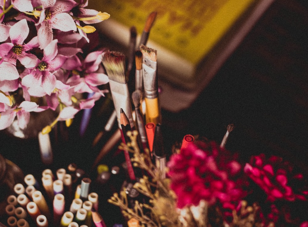 selective focus photography of makeup brush and pencil and pens beside pink and purple flowers