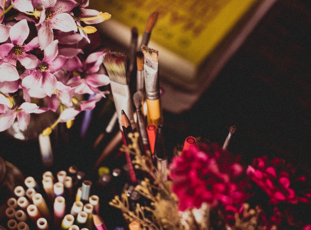 Fotografia selettiva di messa a fuoco del pennello di trucco e della matita e delle penne accanto ai fiori rosa e viola