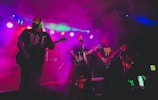 A band performing live on stage, surrounded by colorful lighting and fog. The musicians, each with a guitar, are immersed in their performance with a vibrant display of lights overhead, adding a dynamic and energetic atmosphere.