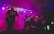 A band performing live on stage, surrounded by colorful lighting and fog. The musicians, each with a guitar, are immersed in their performance with a vibrant display of lights overhead, adding a dynamic and energetic atmosphere.