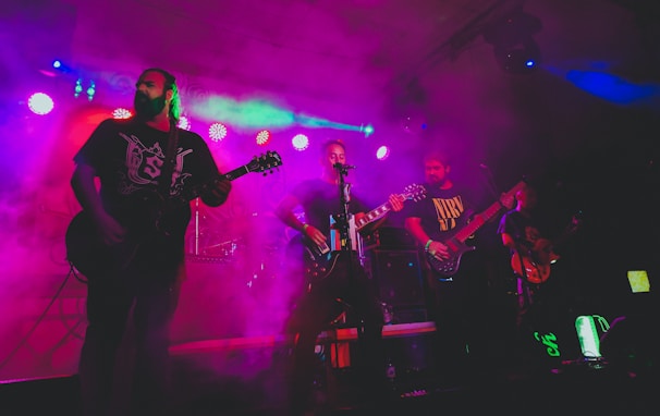 A band performing live on stage, surrounded by colorful lighting and fog. The musicians, each with a guitar, are immersed in their performance with a vibrant display of lights overhead, adding a dynamic and energetic atmosphere.