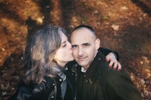 A woman with gray hair kisses a man affectionately on the cheek. They are outdoors on a forest floor, likely during autumn, as the ground is covered with dry leaves. The woman wears a black leather jacket, and the man is dressed in a dark jacket. Their expressions convey warmth and companionship.