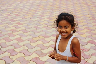 A smiling child holding a handmade drawing, standing in a sunlit village courtyard.