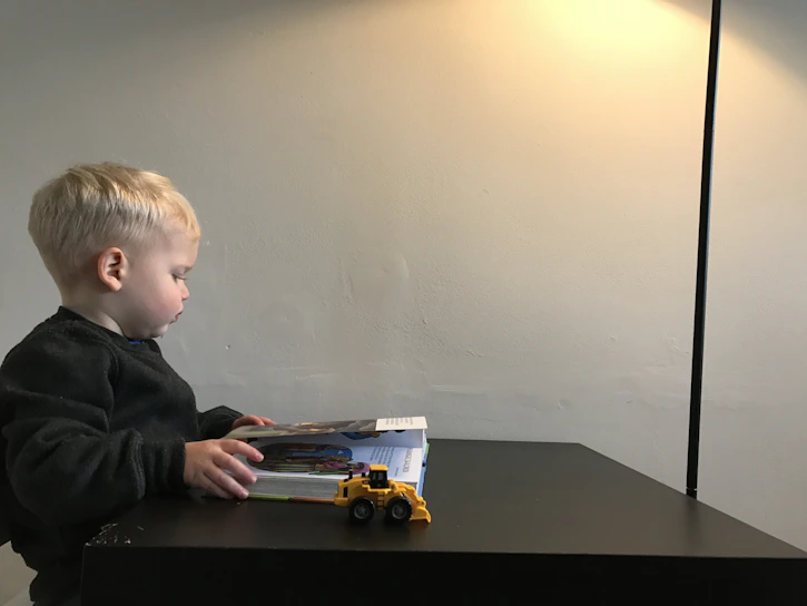 A joyful child sitting calmly and focused while reading a book at a sunny kitchen table.