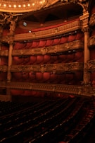 Elegant theater interior with plush red seating and intricate gold architectural details. Multiple balconies are adorned with ornate carvings and soft lighting creates a warm ambiance.