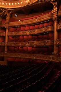 Elegant theater interior with plush red seating and intricate gold architectural details. Multiple balconies are adorned with ornate carvings and soft lighting creates a warm ambiance.