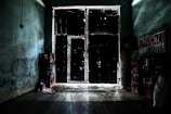 A dimly lit interior features a shadowy, weathered door with small light leaks around its edges. On the left, empty crates are stacked, while on the right, shelves appear to hold additional crates or boxes. A worn sign partially reading 'FARMACIA LOS ANGELES' is visible, adding to the rustic, aged atmosphere.