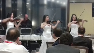 Three women in white dresses are playing violins in what appears to be a formal setting, such as a banquet or reception. An audience is seated in the foreground, and a person is operating sound equipment in the background.