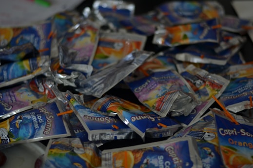 A pile of empty drink pouches of the brand Capri-Sonne, with visible straws attached. The pouches are mostly crumpled and scattered randomly, featuring colorful packaging with blue, orange, and white designs.