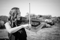 A woman playing a violin outdoors with a serene expression. The scene is in black and white, capturing the elegance and concentration of the musician. The background consists of blurred natural scenery, suggesting a peaceful setting.