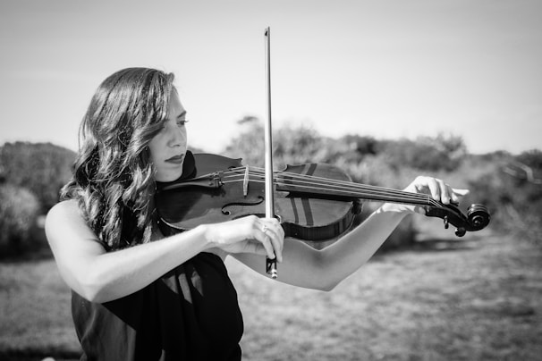 A woman playing a violin outdoors with a serene expression. The scene is in black and white, capturing the elegance and concentration of the musician. The background consists of blurred natural scenery, suggesting a peaceful setting.