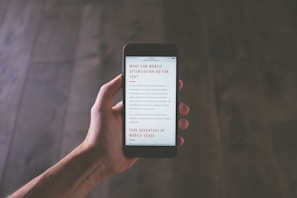 A hand holding a smartphone in a dimly lit environment, displaying text related to mobile optimization. The focus is on the smartphone screen, and the background is a blurred wooden surface.