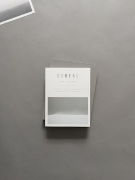 A minimalist magazine cover placed on a flat surface with the title 'Cereal' displayed prominently. The design is simple with a monochrome aesthetic, featuring a small landscape photograph. The backdrop is a muted gray, with a portion of another photograph in the top left corner.