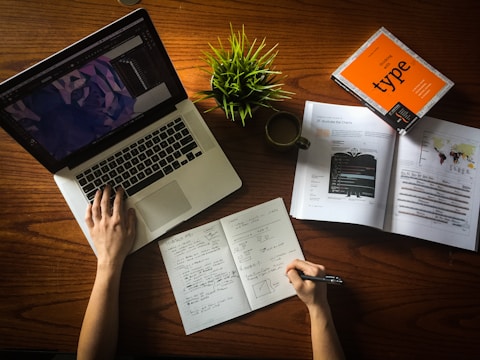 A person is working at a wooden desk with a laptop displaying a design software. Open books and a notebook with handwritten notes are spread out. There is also a potted green plant and a coffee cup on the desk.