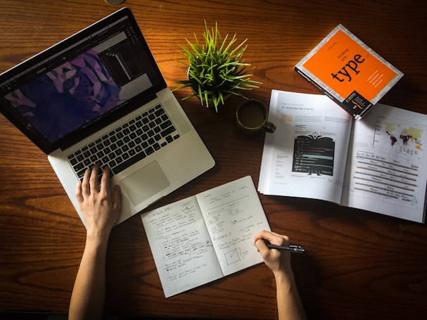 A person is working at a wooden desk with a laptop displaying a design software. Open books and a notebook with handwritten notes are spread out. There is also a potted green plant and a coffee cup on the desk.