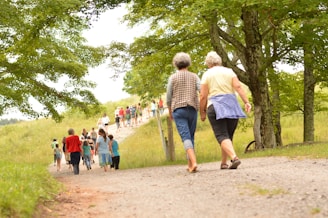 A group of people walks along a gravel path surrounded by lush green scenery. Two older individuals are prominently seen in the foreground, walking together along the path. The area is dotted with trees and grassy fields, creating a serene and natural environment.