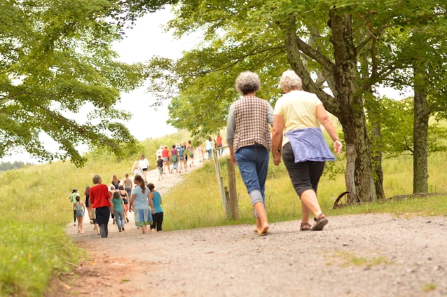 A group of people walks along a gravel path surrounded by lush green scenery. Two older individuals are prominently seen in the foreground, walking together along the path. The area is dotted with trees and grassy fields, creating a serene and natural environment.