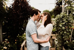 A couple is standing in a lush greenhouse surrounded by various plants. The woman is wearing a white blouse and jeans, while the man is dressed in a light denim shirt. They are facing each other with loving expressions, suggesting a romantic connection.