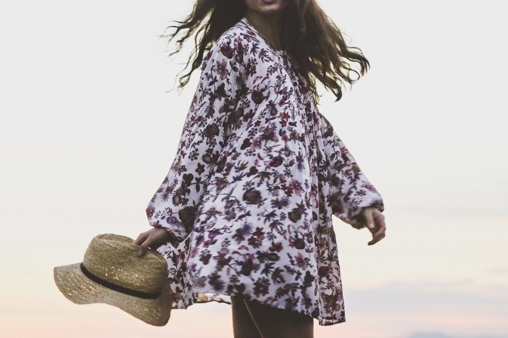 Mujer que viste camisa de manga larga floral blanca y marrón mientras sostiene un sombrero fedora marrón