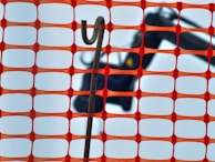 An orange construction fence with a rectangular grid pattern is in the foreground. Behind the fence, a hooked metal rod is visible against a blurred background that appears to include construction equipment.