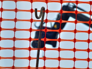 An orange construction fence with a rectangular grid pattern is in the foreground. Behind the fence, a hooked metal rod is visible against a blurred background that appears to include construction equipment.