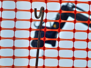 An orange construction fence with a rectangular grid pattern is in the foreground. Behind the fence, a hooked metal rod is visible against a blurred background that appears to include construction equipment.