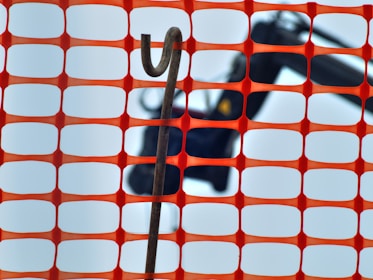 An orange construction fence with a rectangular grid pattern is in the foreground. Behind the fence, a hooked metal rod is visible against a blurred background that appears to include construction equipment.