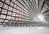 An intricate lattice of metallic mesh forms a tunnel-like structure, with interwoven wires creating an abstract pattern. Some wires are highlighted in red, creating a contrast against the predominant silver. The mesh appears to extend and twist, forming a dynamic, curved form.
