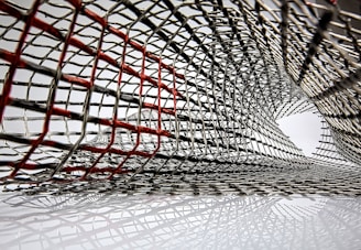 An intricate lattice of metallic mesh forms a tunnel-like structure, with interwoven wires creating an abstract pattern. Some wires are highlighted in red, creating a contrast against the predominant silver. The mesh appears to extend and twist, forming a dynamic, curved form.
