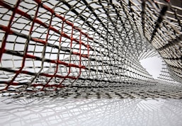 An intricate lattice of metallic mesh forms a tunnel-like structure, with interwoven wires creating an abstract pattern. Some wires are highlighted in red, creating a contrast against the predominant silver. The mesh appears to extend and twist, forming a dynamic, curved form.