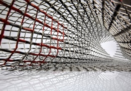 An intricate lattice of metallic mesh forms a tunnel-like structure, with interwoven wires creating an abstract pattern. Some wires are highlighted in red, creating a contrast against the predominant silver. The mesh appears to extend and twist, forming a dynamic, curved form.