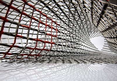 An intricate lattice of metallic mesh forms a tunnel-like structure, with interwoven wires creating an abstract pattern. Some wires are highlighted in red, creating a contrast against the predominant silver. The mesh appears to extend and twist, forming a dynamic, curved form.