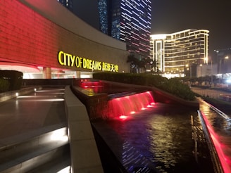 A nighttime urban scene with the City of Dreams building prominently displayed. The building features illuminated signage in yellow characters alongside red lighting. In the foreground, there is a water feature with cascading water highlighted by red lights, surrounded by walkways and lush greenery. The background showcases tall buildings with modern, lit-up facades, adding to the vibrant city atmosphere.