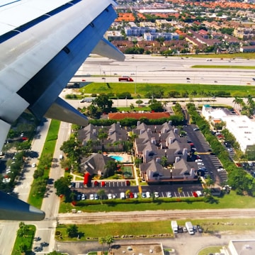This image captures an aerial view from an airplane window. The visible airplane wing suggests the aircraft is either taking off or landing. Below, there is a residential complex with neatly arranged houses and a central swimming pool. Surrounding the residential area are roads with moving cars, green spaces, parking lots, and nearby buildings. In the distance, a highway with vehicles, and a backdrop of further urban development can be seen.